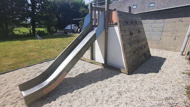 Une aire de jeux avec un toboggan et un mur d’escalade, entourée d’une zone recouverte de gravier et d’une pelouse.