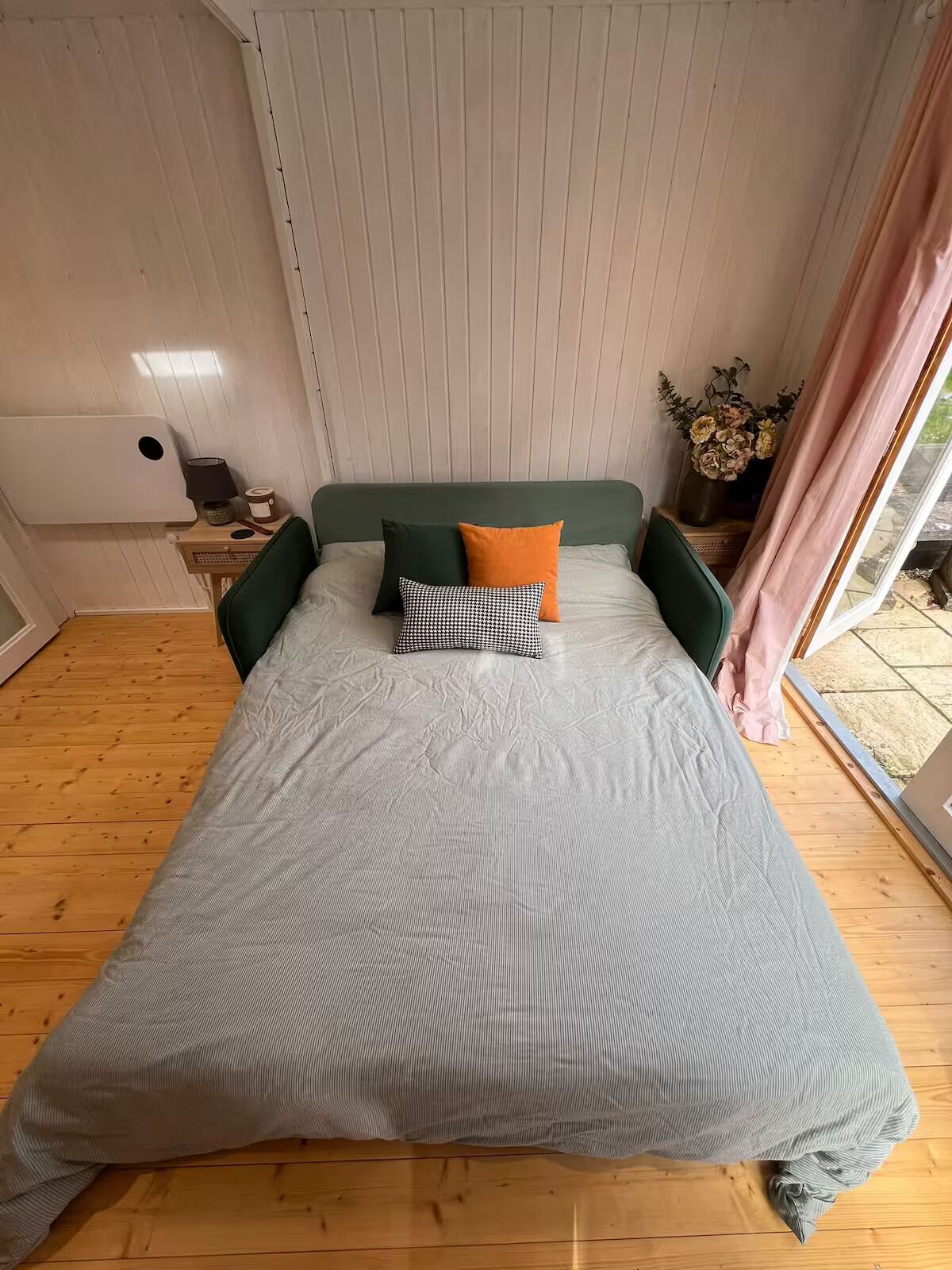 Ein Bett mit grauer Streifenoptik, einem grünen Kopfteil und zwei Kissen – eines kariert in Schwarz und Weiß, das andere orange. Neben dem Bett steht ein kleiner Tisch mit einer Lampe und einem Blumenstrauß.