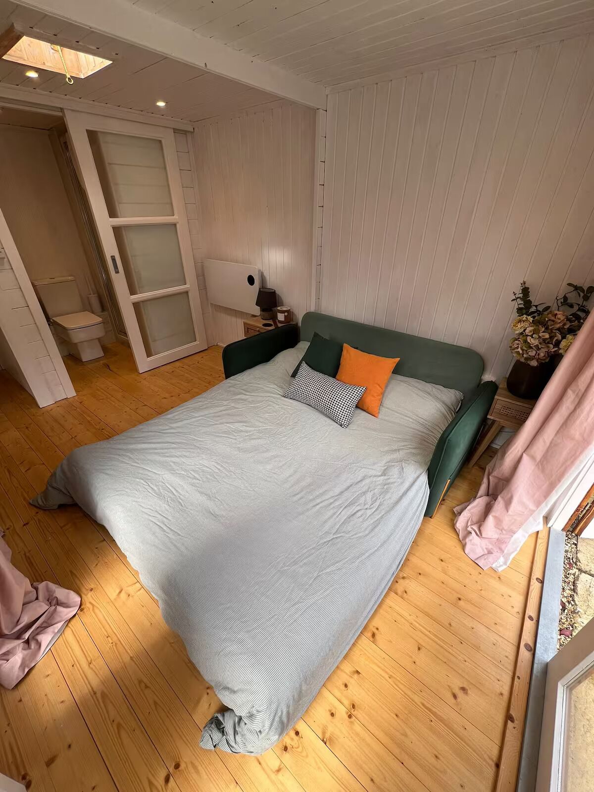A bedroom with a bed, wooden flooring, a nightstand, a lamp, and a window with curtains.