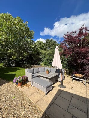 Een patio met een grijze modulaire bank, een ronde tafel en een roze parasol. Achtergrond met potplanten en een vuurkorf.