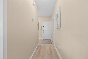Un pasillo con paredes color beige, piso de madera, una puerta blanca y un tapete pequeño en la entrada.