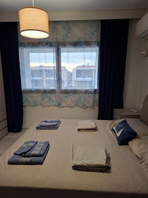Une chambre d’hôtel comprenant un lit, une table de chevet, une lampe, une fenêtre avec des rideaux et un appareil de climatisation.