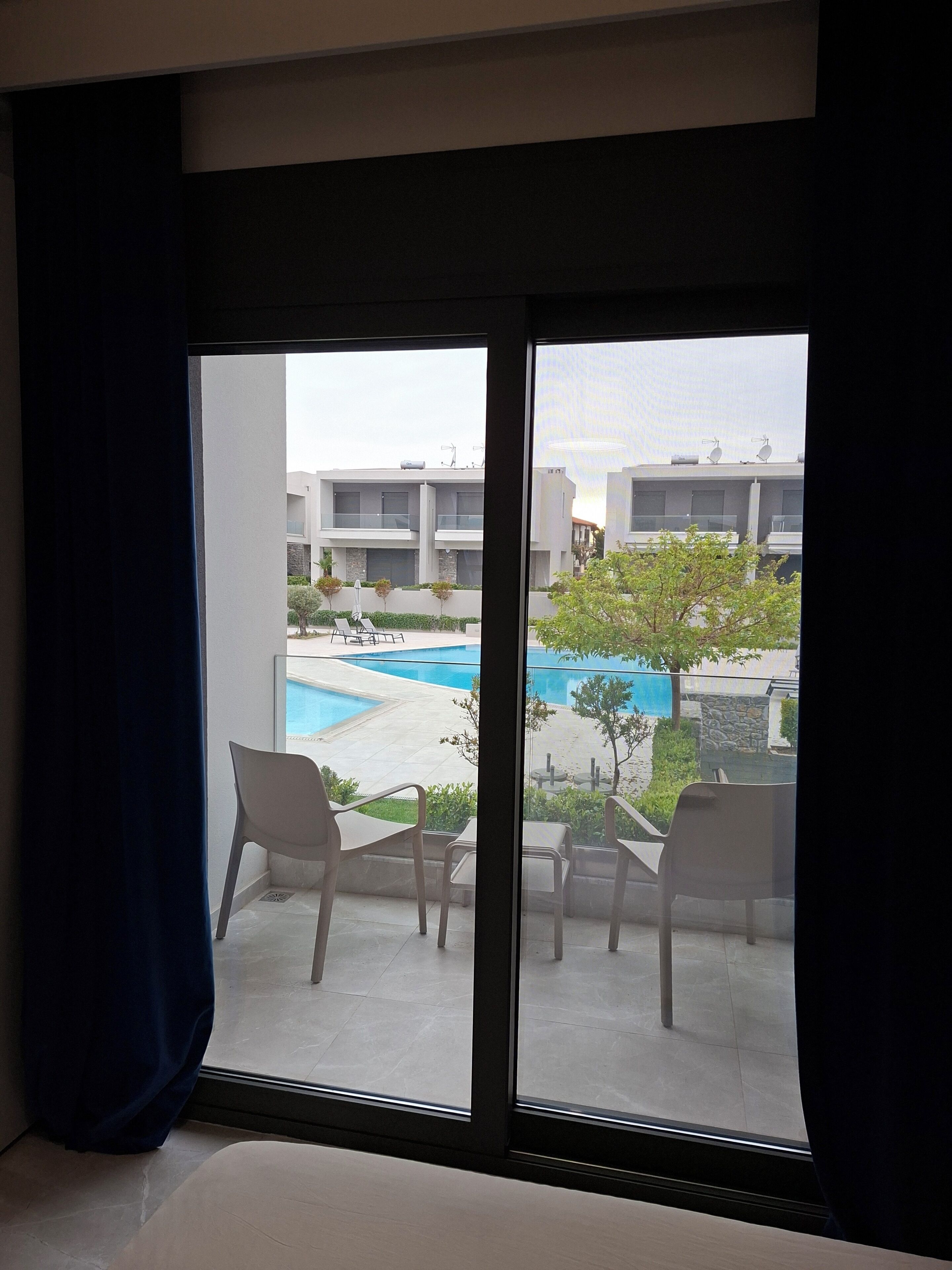 Quarto com uma grande porta de vidro deslizante que dá para área externa com piscina, cadeiras e vista de edifícios modernos.