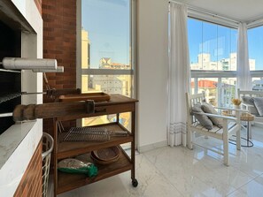 A modern apartment with a kitchen area featuring a wooden cart, a large window with a city view, and a balcony with outdoor seating. - Brisa de Palmas - Res. Maria Eduarda (Governador Celso Ramos)
