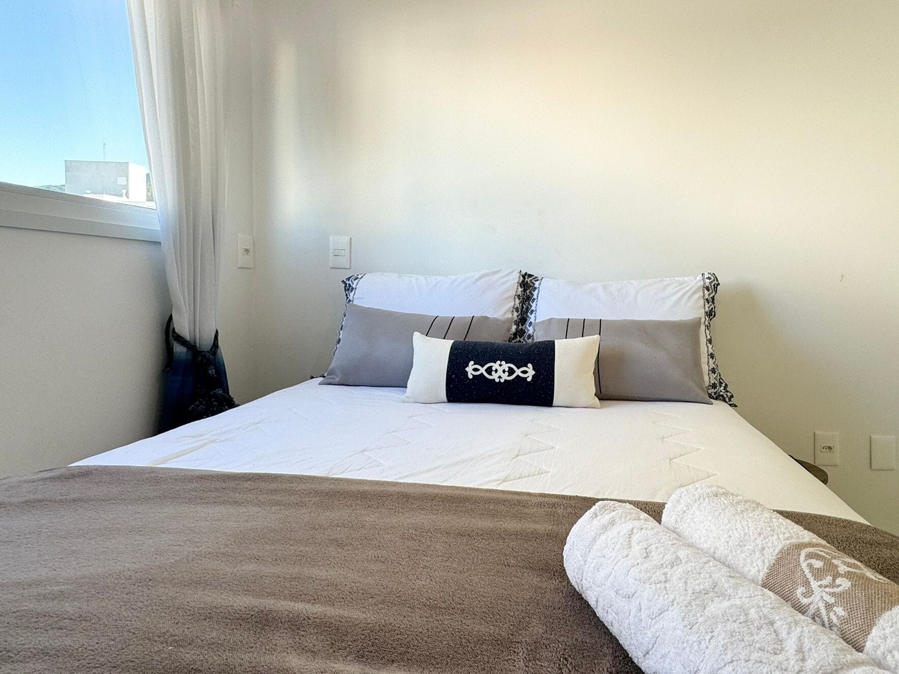 A neatly made bed with a brown blanket and pillows, a window with curtains, and a white wall.