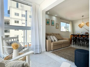 A modern living room with a sofa, dining table, and chairs. There is a window with curtains, a view of buildings outside, and a ceiling light fixture. - Brisa de Palmas - Res. Maria Eduarda (Governador Celso Ramos)