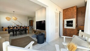A modern living room with a dining area, a TV mounted on a brick wall, and a white sofa. - Brisa de Palmas - Res. Maria Eduarda (Governador Celso Ramos)