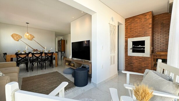 A modern living room with a dining area, a TV mounted on a brick wall, and a white sofa. - Brisa de Palmas - Res. Maria Eduarda (Governador Celso Ramos)