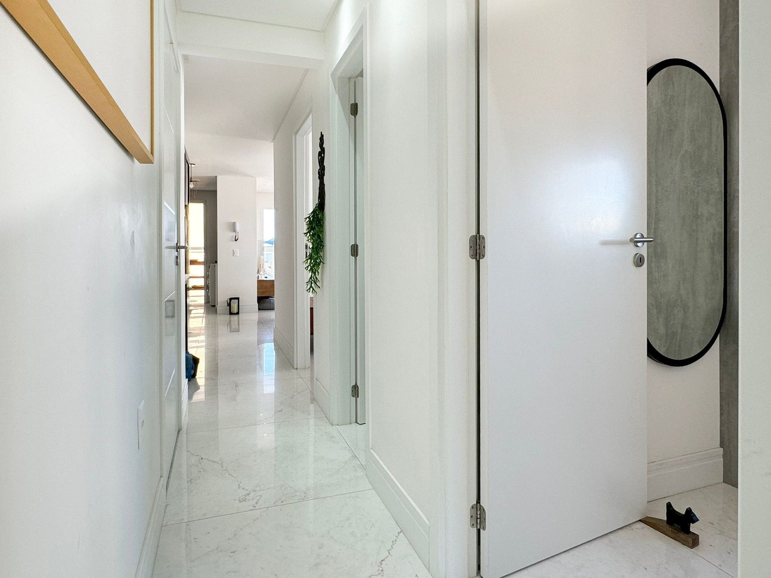 A hallway with white walls and a marble floor, leading to a room with a door that has a circular mirror and a small plant.