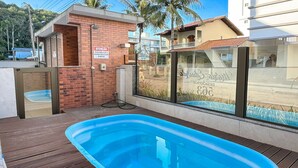 A swimming pool with a wooden deck, a small brick structure, and a glass window with a reflection. - Brisa de Palmas - Res. Maria Eduarda (Governador Celso Ramos)