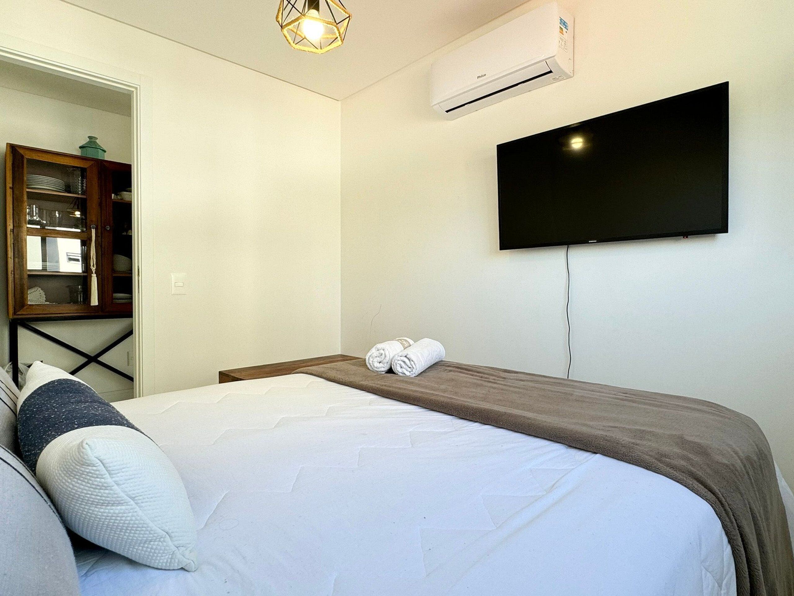 A hotel room with a bed, a flat-screen TV mounted on the wall, an air conditioning unit, and a wooden cabinet with glass doors.