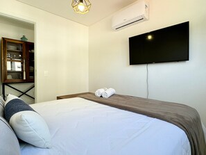 Habitación de hotel con cama, televisor de pantalla plana montado en la pared, unidad de aire acondicionado y mueble de madera con puertas de cristal.
