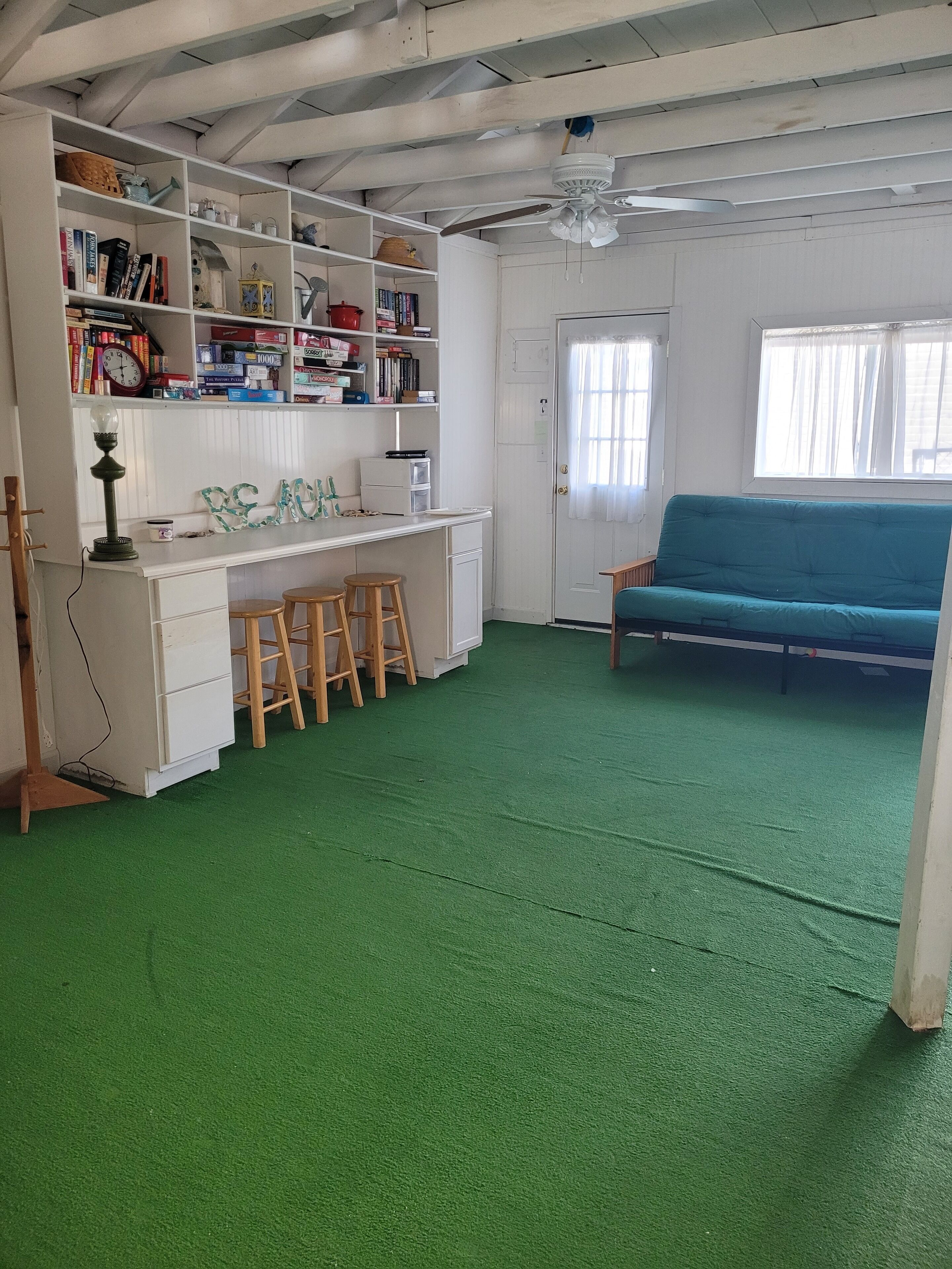 Ett rum med en grön matta, vita väggar, en blå soffa, ett vitt skrivbord med böcker och en lampa samt en takfläkt.