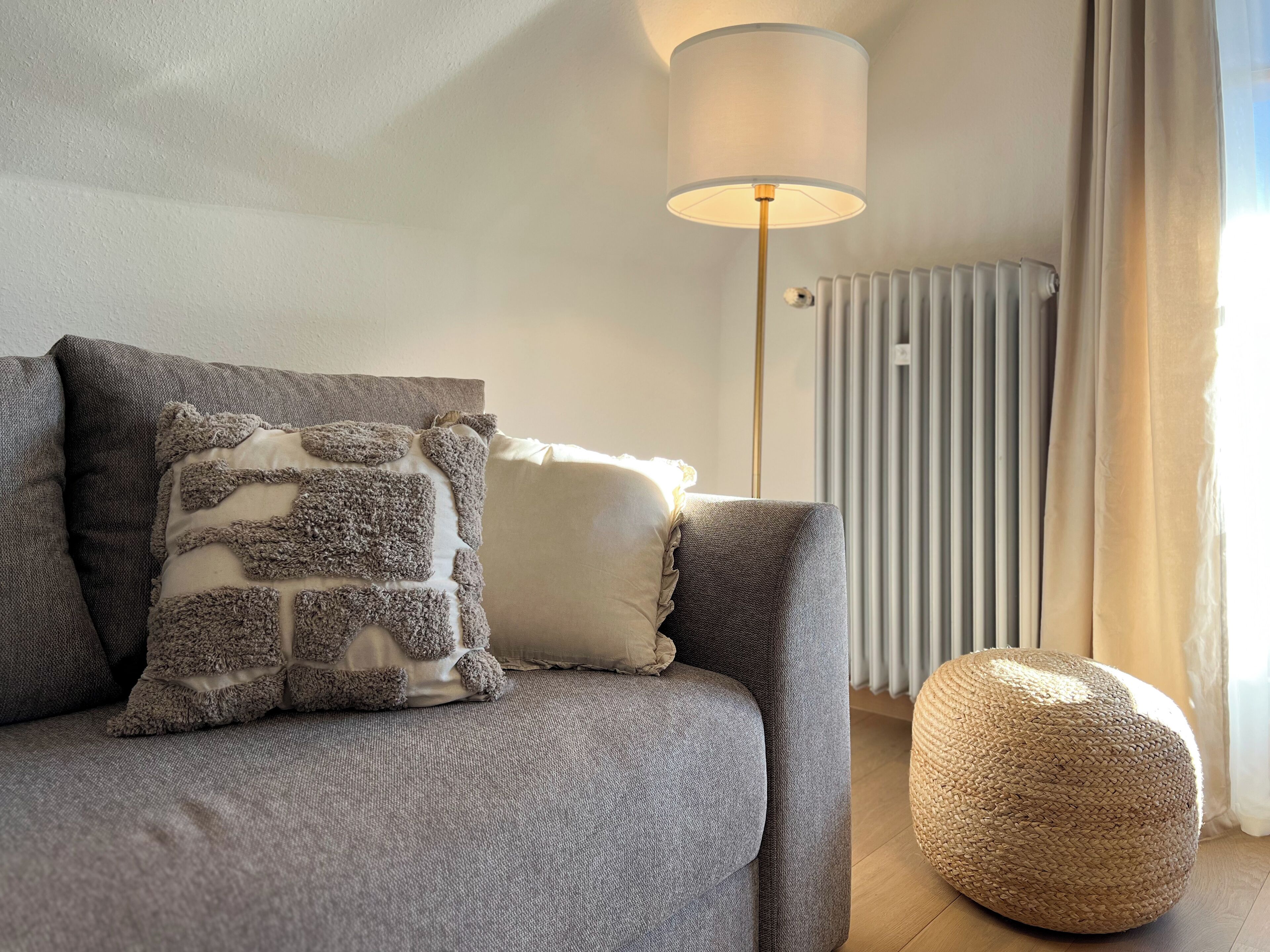 Un salon moderne avec un canapé gris, un coussin décoratif à texture, une lampe sur pied, un radiateur et un pouf tressé.