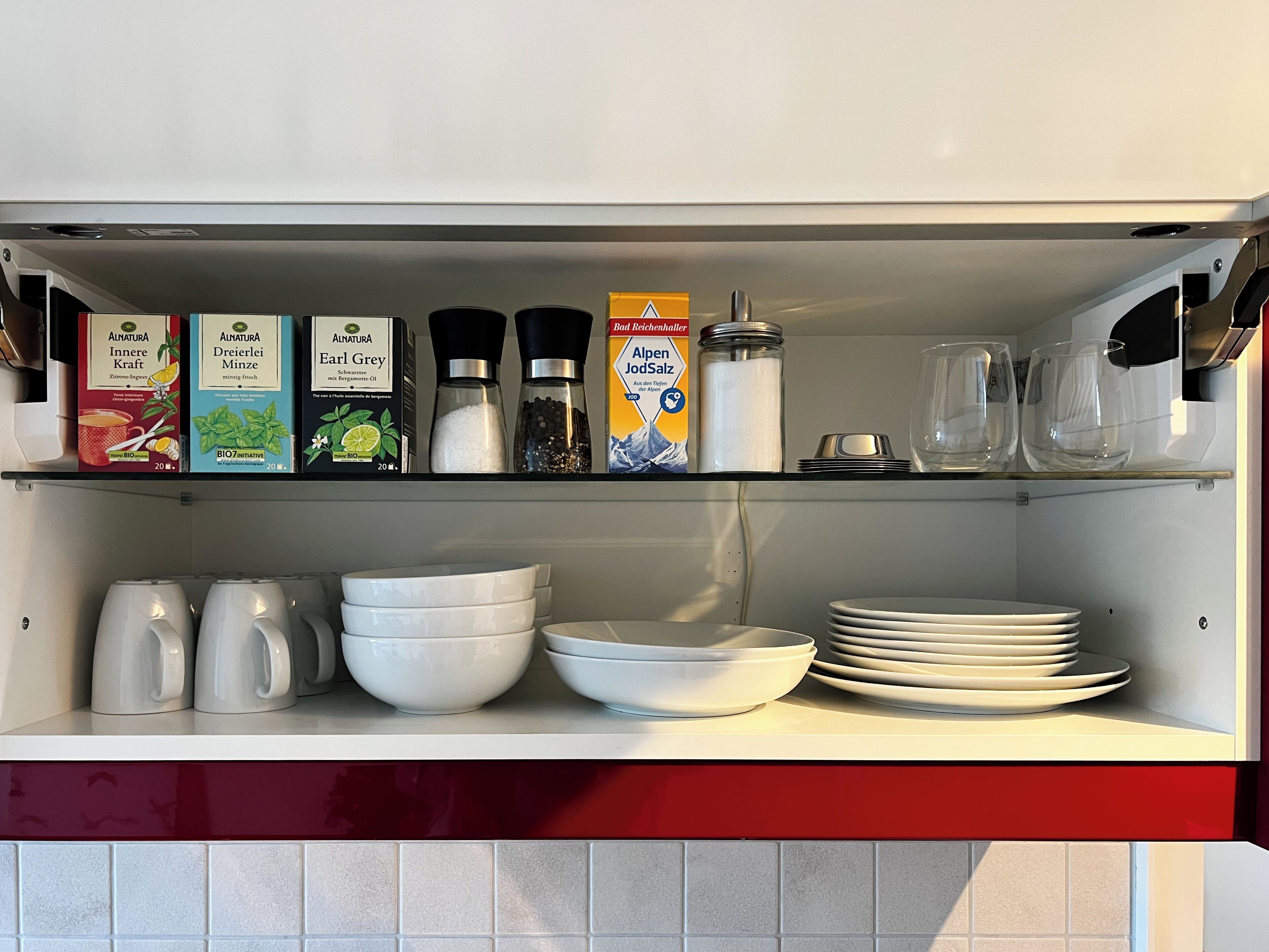 A kitchen shelf with various items including boxes of Alnatura products, salt and pepper shakers, a jar of salt, and stacked white bowls and plates.