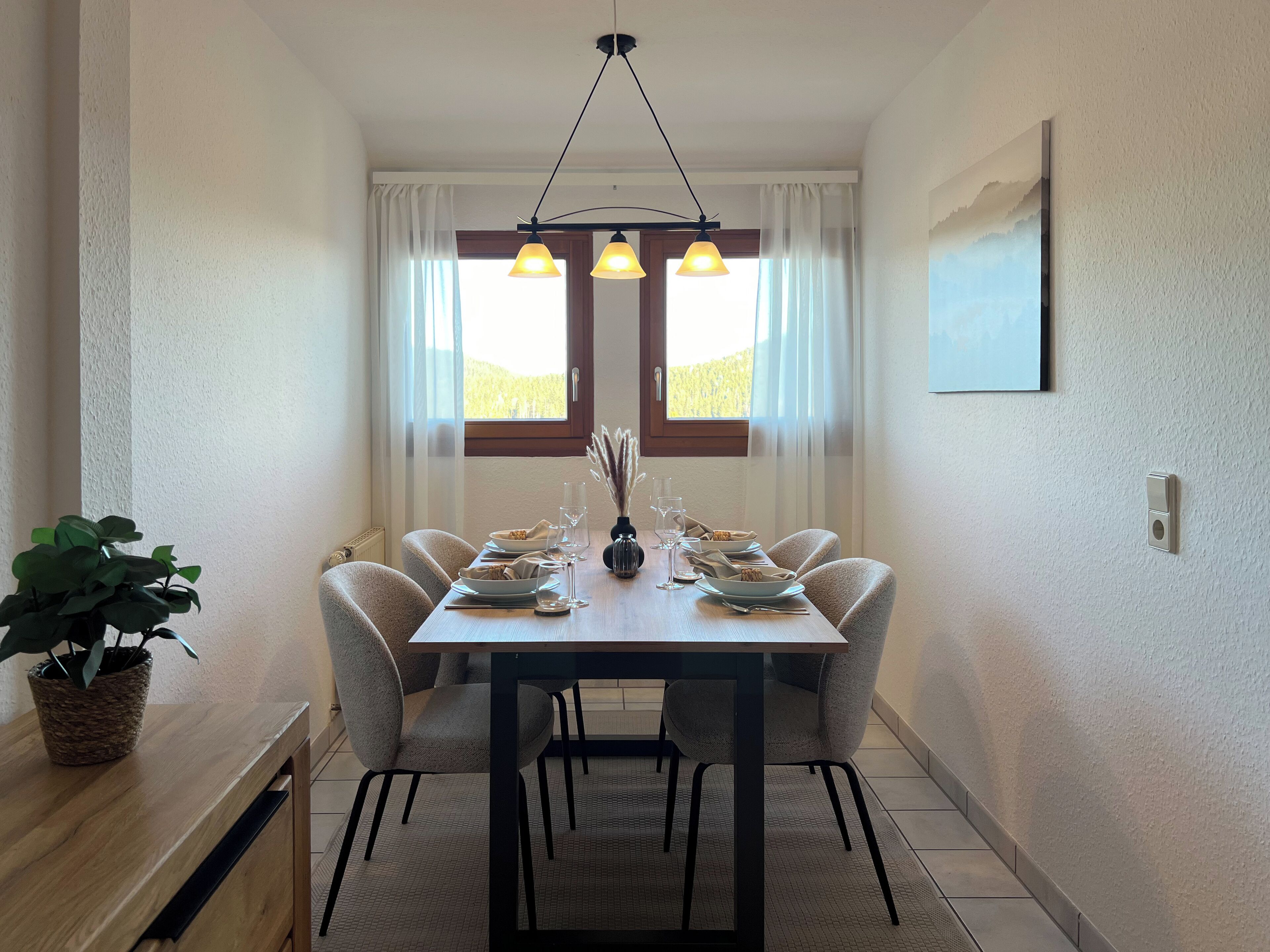 A dining area with a wooden table, four chairs, a potted plant, and a pendant light fixture.