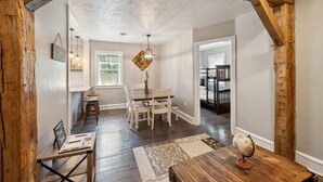 Un coin repas avec une table et des chaises en bois, un lustre et une vue sur une chambre avec des lits superposés.