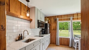 Une cuisine moderne avec des meubles blancs, un évier en acier inoxydable et un four à micro-ondes. Une fenêtre offre une vue sur la verdure environnante.