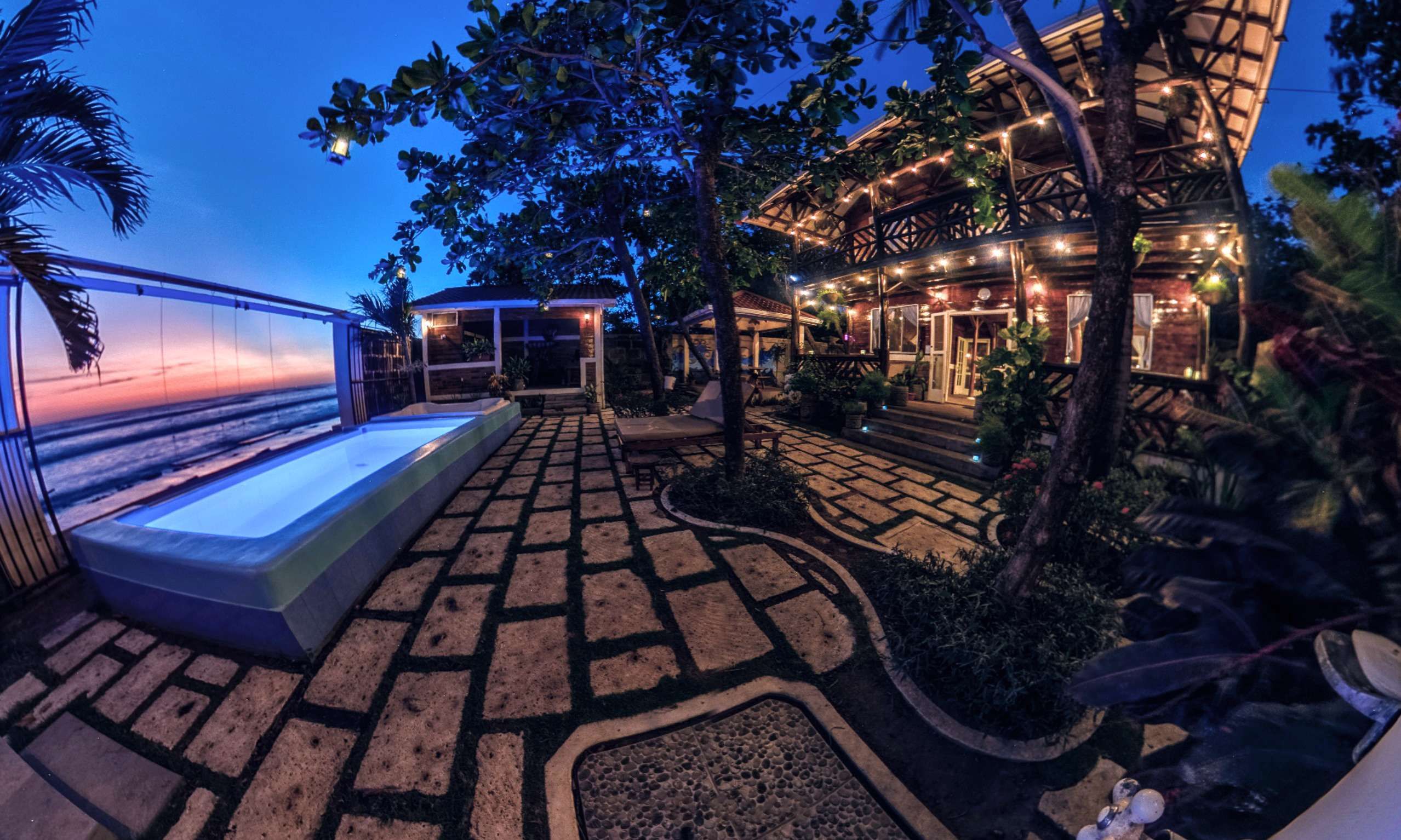 A pool area with a view of the ocean, a covered seating area, and a building with string lights.