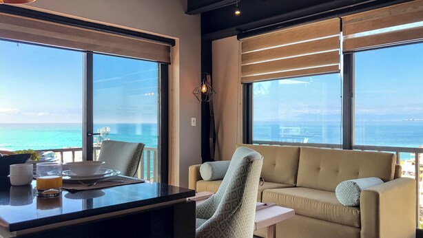 A modern room with a large window overlooking the sea, a sofa, a chair, and a small table with breakfast items. - Los Muertos Beach Walking Distance Suite - C406 (Puerto Vallarta)