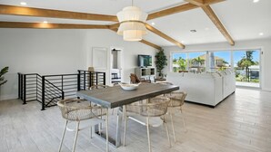 Een ruime woonkamer met een eettafel, stoelen en een bank. Er is een balkon met uitzicht op palmbomen en een huis.