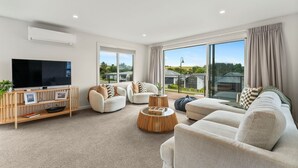 Una sala de estar moderna con un televisor de pantalla plana, asientos cómodos, un mueble de entretenimiento de madera y ventanas grandes que ofrecen vistas al exterior.