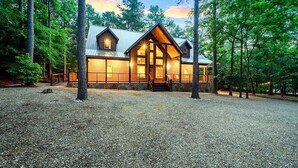A rustic cabin with a stone facade, large windows, and a metal roof, surrounded by tall trees. - Simmer Down: Shuffleboard, Pool Table, Hot Tub, Firepit, Sleeps 17 (Broken Bow)