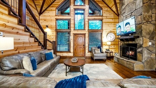 A log cabin interior with a stone fireplace, leather sofas, a wooden staircase, and a large window overlooking the forest. - Simmer Down: Shuffleboard, Pool Table, Hot Tub, Firepit, Sleeps 17 (Broken Bow)