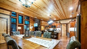 A wooden interior with a dining table, chairs, a television, and a kitchen area. - Simmer Down: Shuffleboard, Pool Table, Hot Tub, Firepit, Sleeps 17 (Broken Bow)