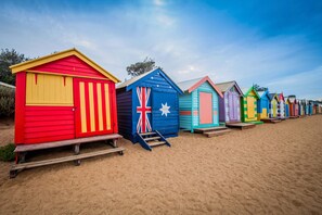A row of colorful beach huts with steps leading up to them, set on a sandy beach. - Artist Haven With Parking By My Getaways (Brighton)