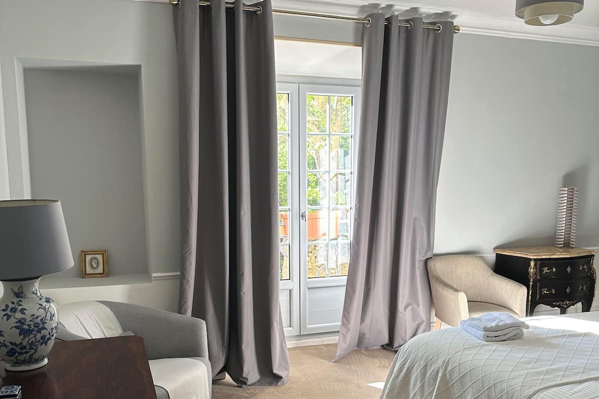 Une chambre à coucher comprenant un lit, une chaise, une table de chevet, une lampe et une fenêtre avec des rideaux, offrant une vue sur l’extérieur.