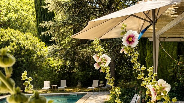Ett område vid poolen med en stenbelagd uteplats, solstolar, en trätunna och en pergola med ett parasoll.