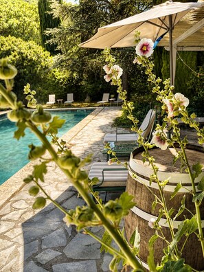Uma área à beira da piscina com um terraço de pedra, cadeiras de descanso, um barril de madeira e uma pérgula com guarda-sol.