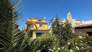 A yellow building with a red-tiled roof, surrounded by lush greenery and flowering plants. - Relaxing 5-Bedroom Villa with Private Pool, Outdoor Playground, Barbeque zona.

 (Carcavelos)