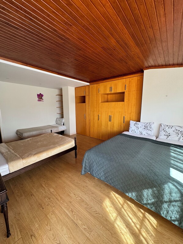 Un dormitorio con techo de madera, una cama con colcha azul, un armario de madera y una pequeña mesa con una silla.