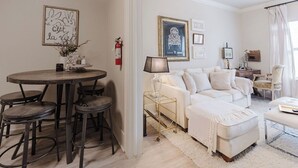 Ein modernes Wohnzimmer mit einem weißen Sofa, einem runden Esstisch mit Hocker und einem Couchtisch. An den Wänden hängen gerahmte Bilder und ein Feuerlöscher ist angebracht.