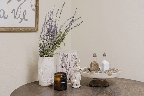 Une table ronde en bois sur laquelle se trouve un vase en céramique blanche contenant de la lavande, une œuvre d’art encadrée représentant un lapin, une petite figurine de lapin en céramique, et deux bouteilles en verre contenant des salières et des poivrières.