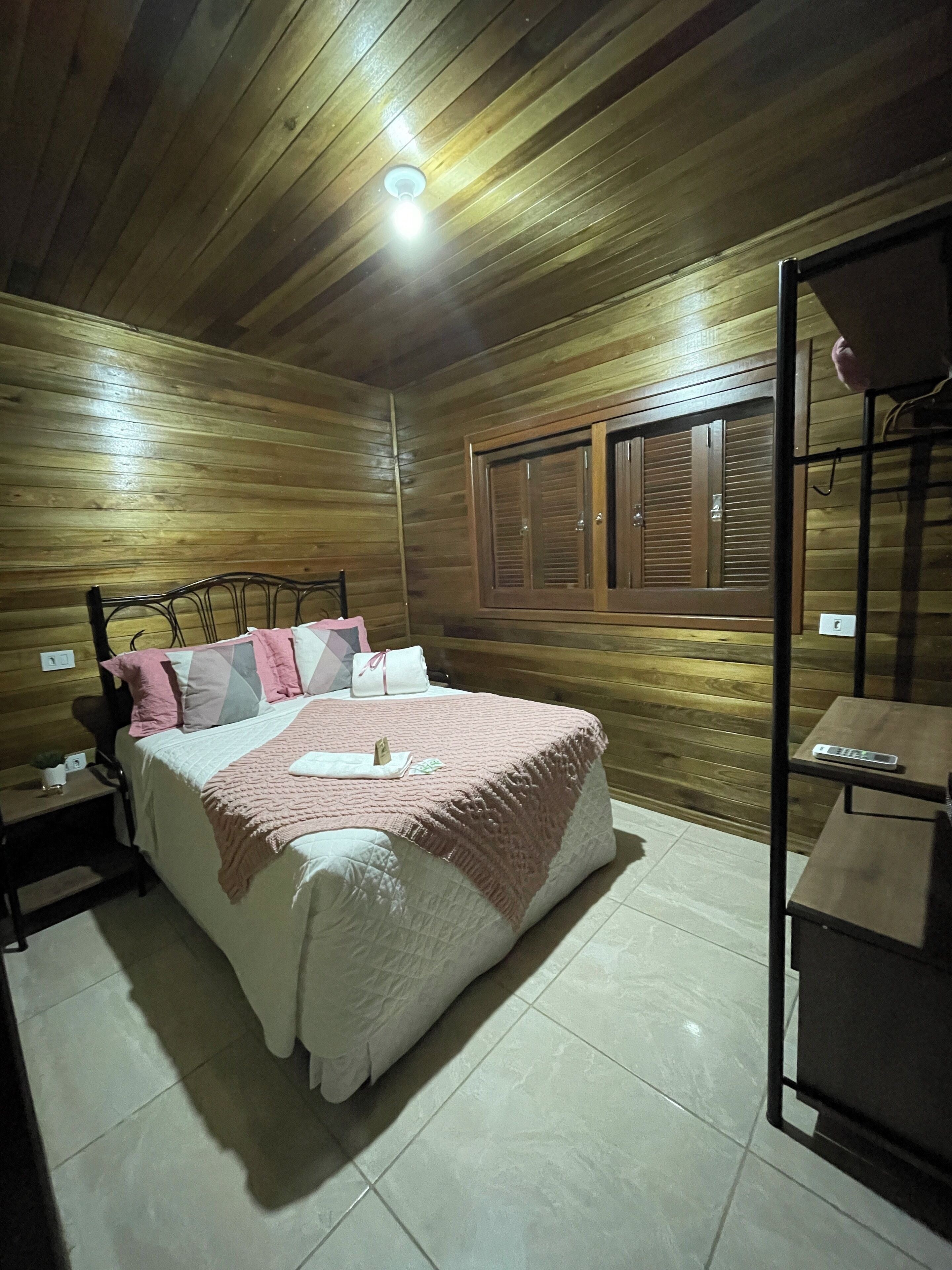 Une chambre à coucher aux murs lambrissés, avec un lit, des tables de chevet, une fenêtre avec des volets et un luminaire au plafond.