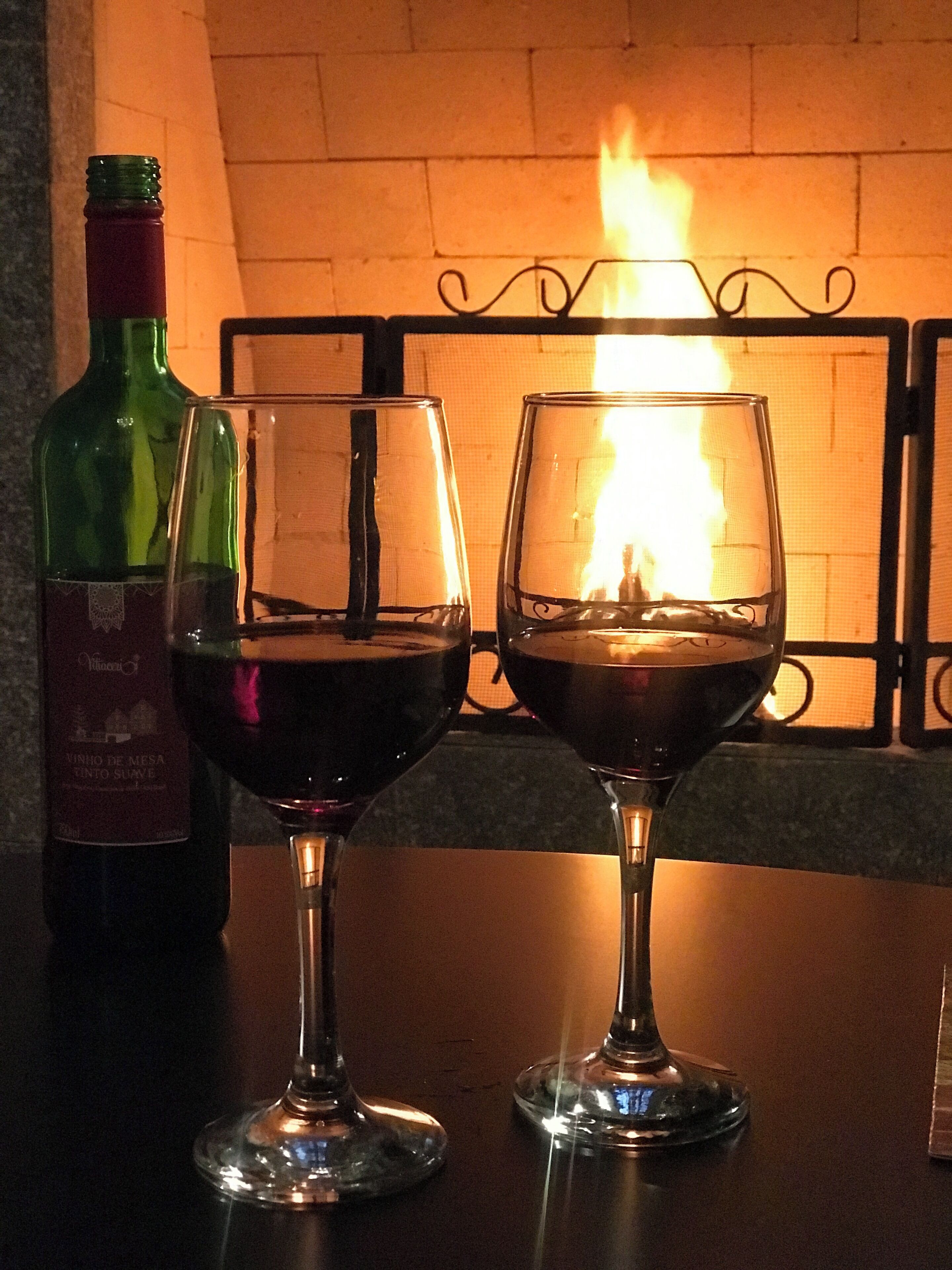 Dos copas de vino sobre una mesa, con una botella y una chimenea al fondo.