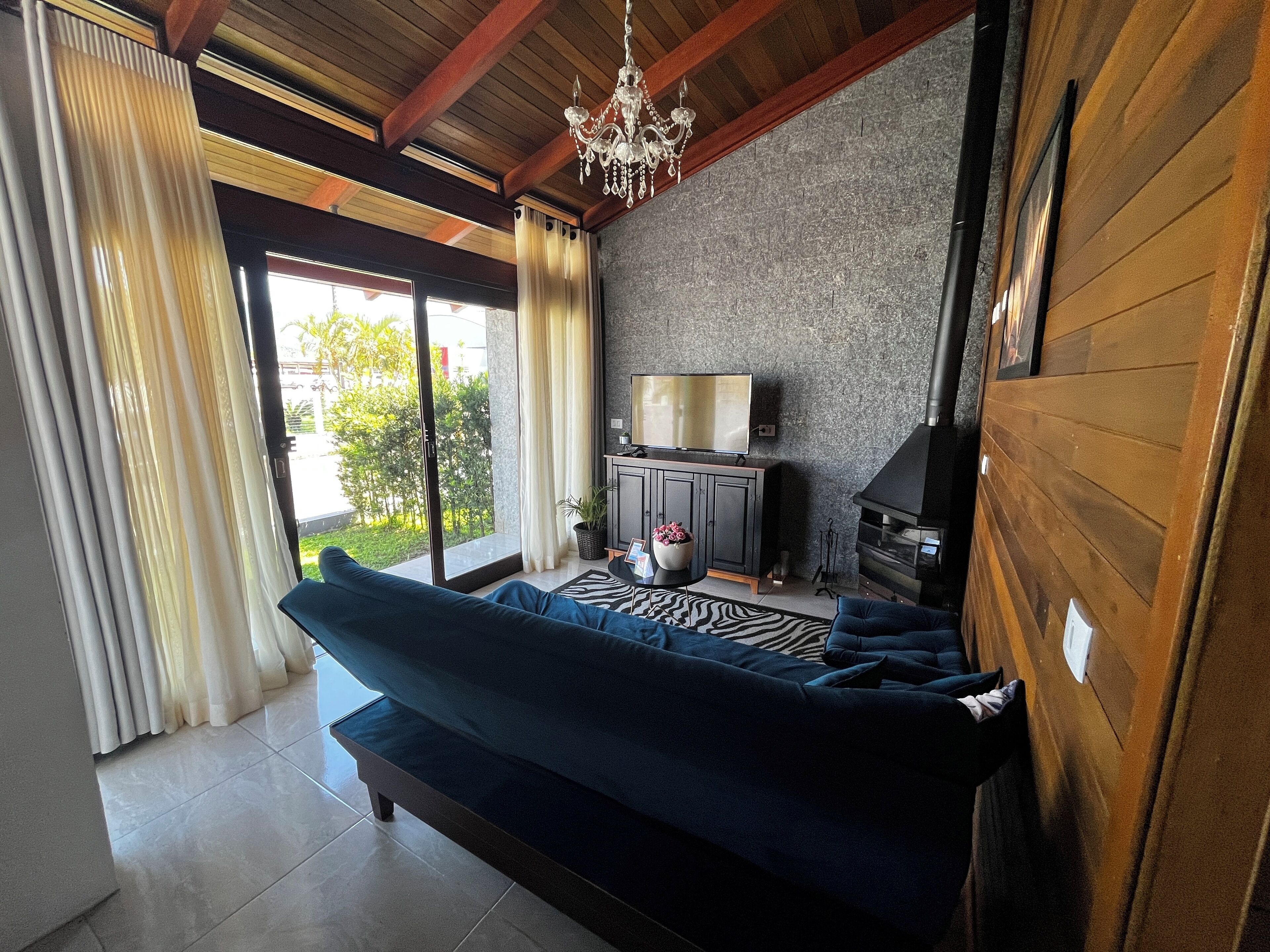 A modern living room with a dark blue sofa, a television on a wooden cabinet, a fireplace, and a view of greenery outside.