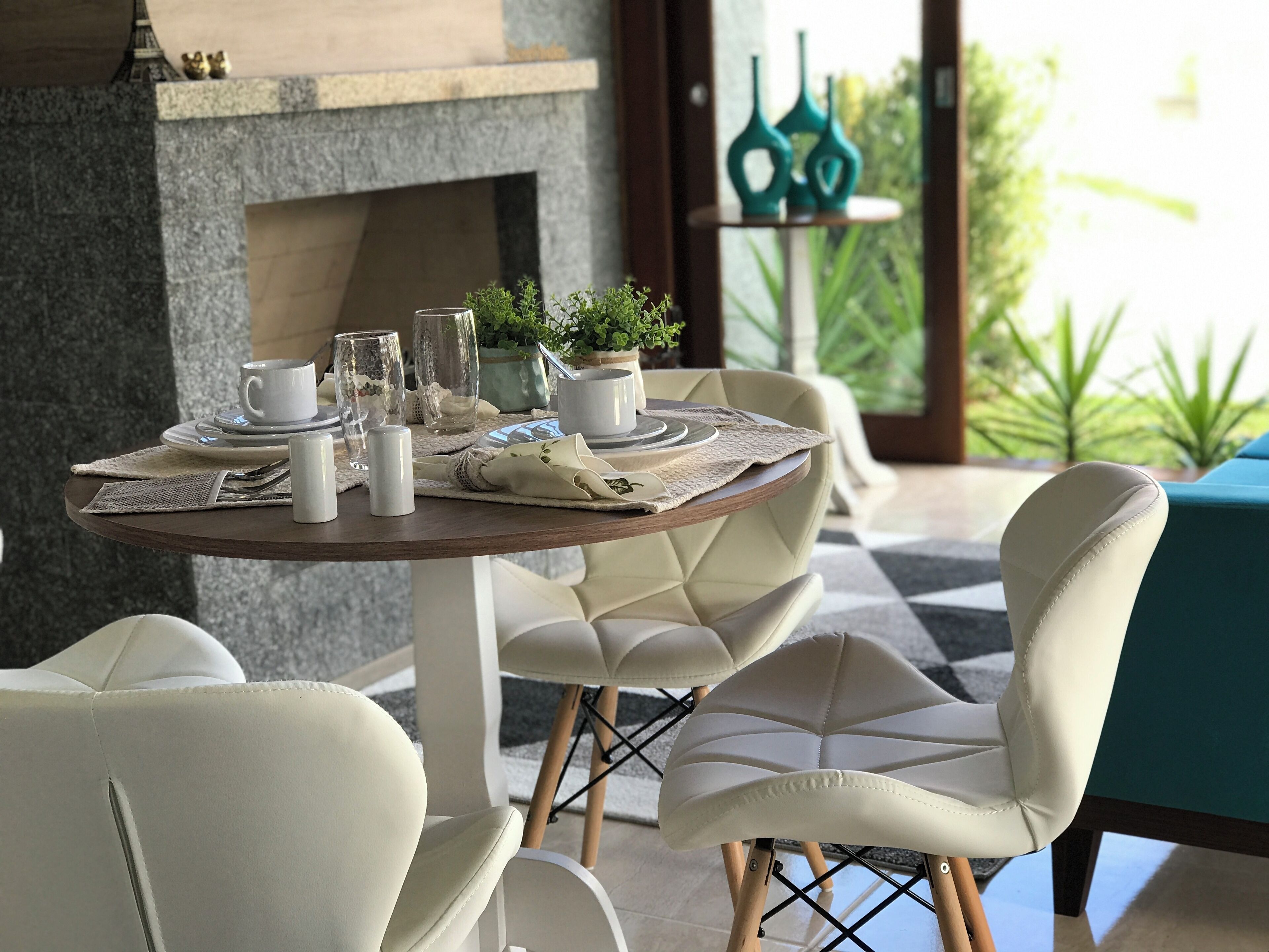 A dining area with a round table set for two, surrounded by white upholstered chairs. A fireplace is visible in the background, and there is a view of greenery through the window.