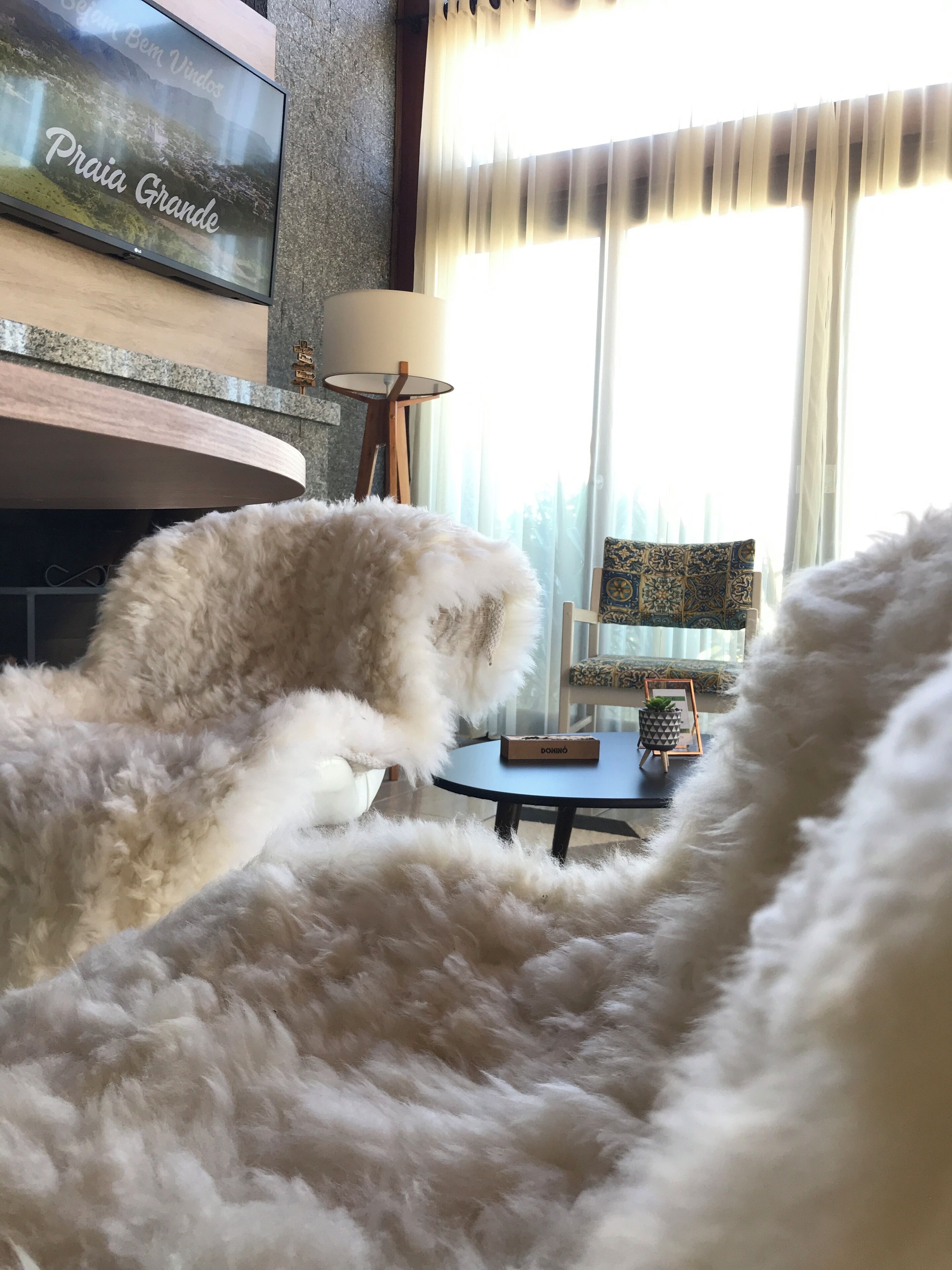 Una habitación con un alfombra blanca y mullida, un televisor montado en una pared de piedra, una pequeña mesa redonda con una caja encima y una ventana con cortinas translúcidas.