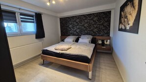 A bedroom with a bed, a radiator, a window with blinds, a wall with a floral pattern, and a framed picture on the wall. - Eifelglück (Hirten)