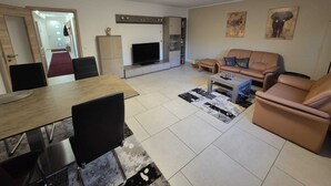 A living room with a TV, sofa, coffee table, and dining area with chairs. - Eifelglück (Hirten)