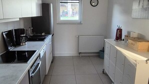 A compact kitchen with a stove, oven, and sink. There's a window with a view of the outside, a radiator, and a clock on the wall. - Eifelglück (Hirten)
