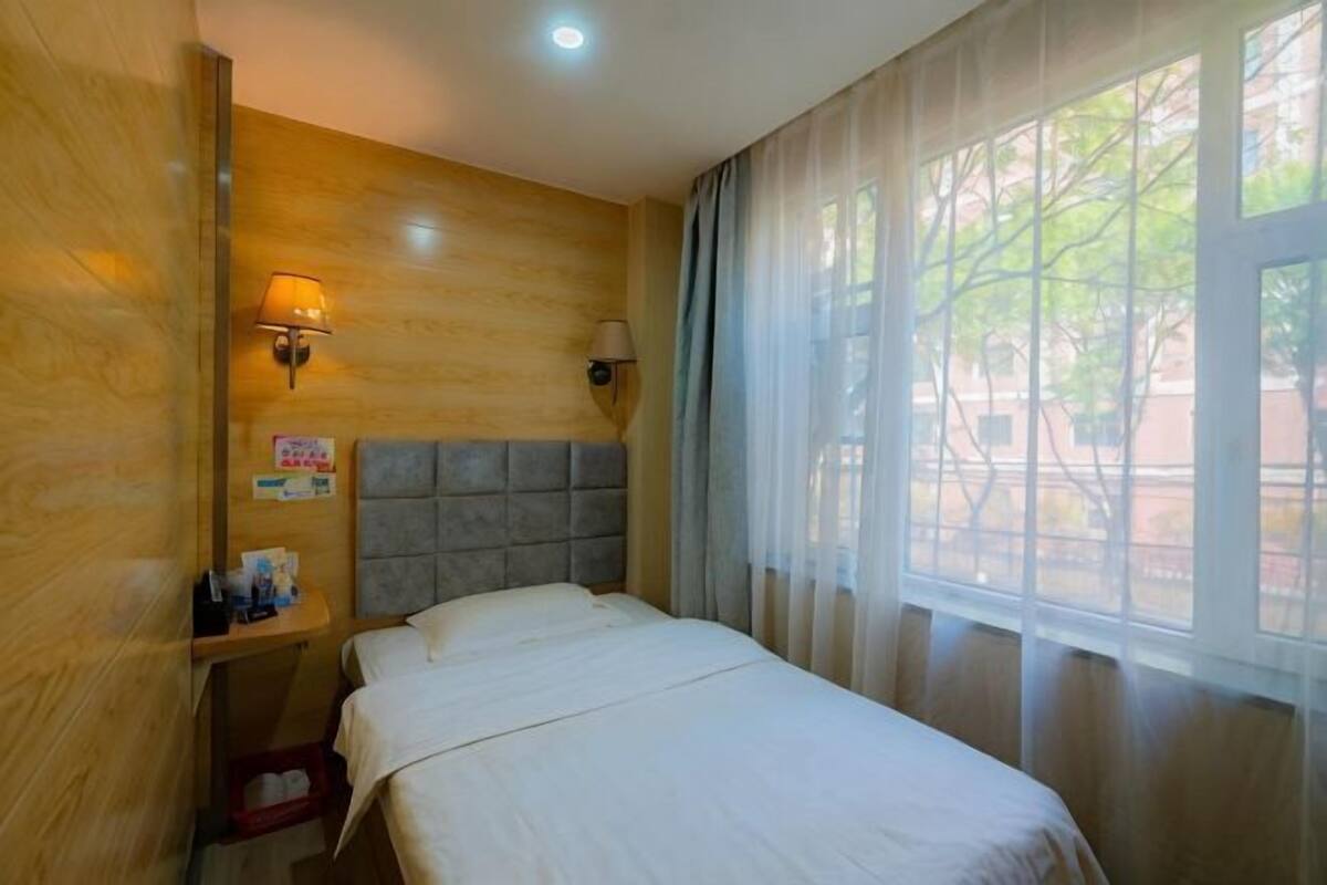 A hotel room with a bed, a nightstand, a wall-mounted lamp, and a window with curtains.