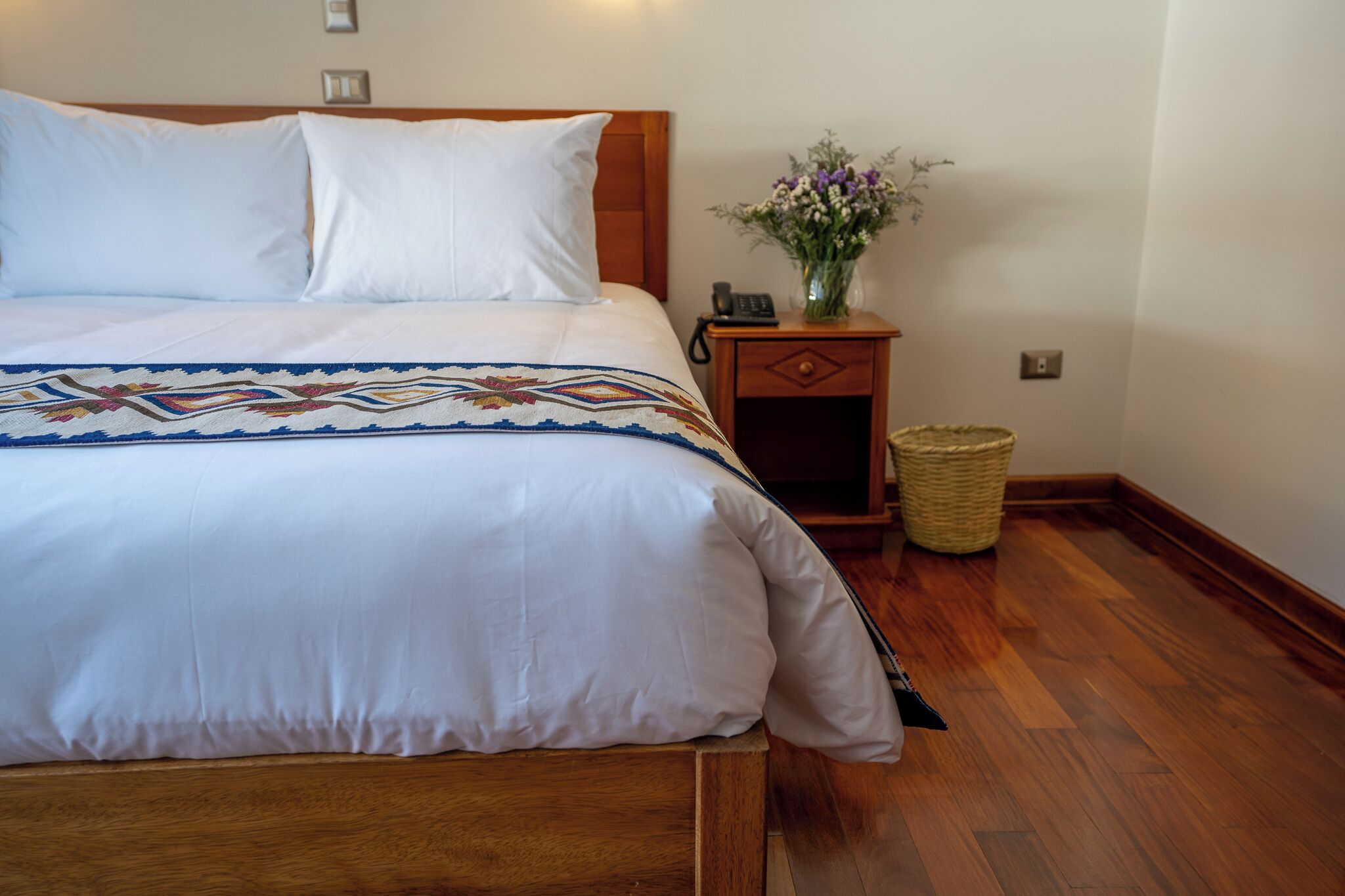 Una cama bien tendida con una colcha estampada, un cabecero de madera, una mesita de noche con un jarrón de flores y una cesta de mimbre.