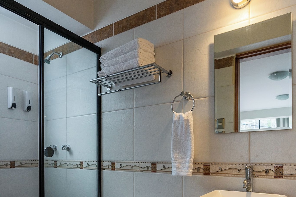 Un baño moderno con ducha de vidrio, inodoro blanco, lavamanos con espejo y toallero con toallas dobladas.