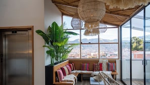 A modern interior with a large window, a wooden bench with colorful cushions, a wicker chair, and a plant. - Cavana Hotel Boutique Cusco (Cusco)