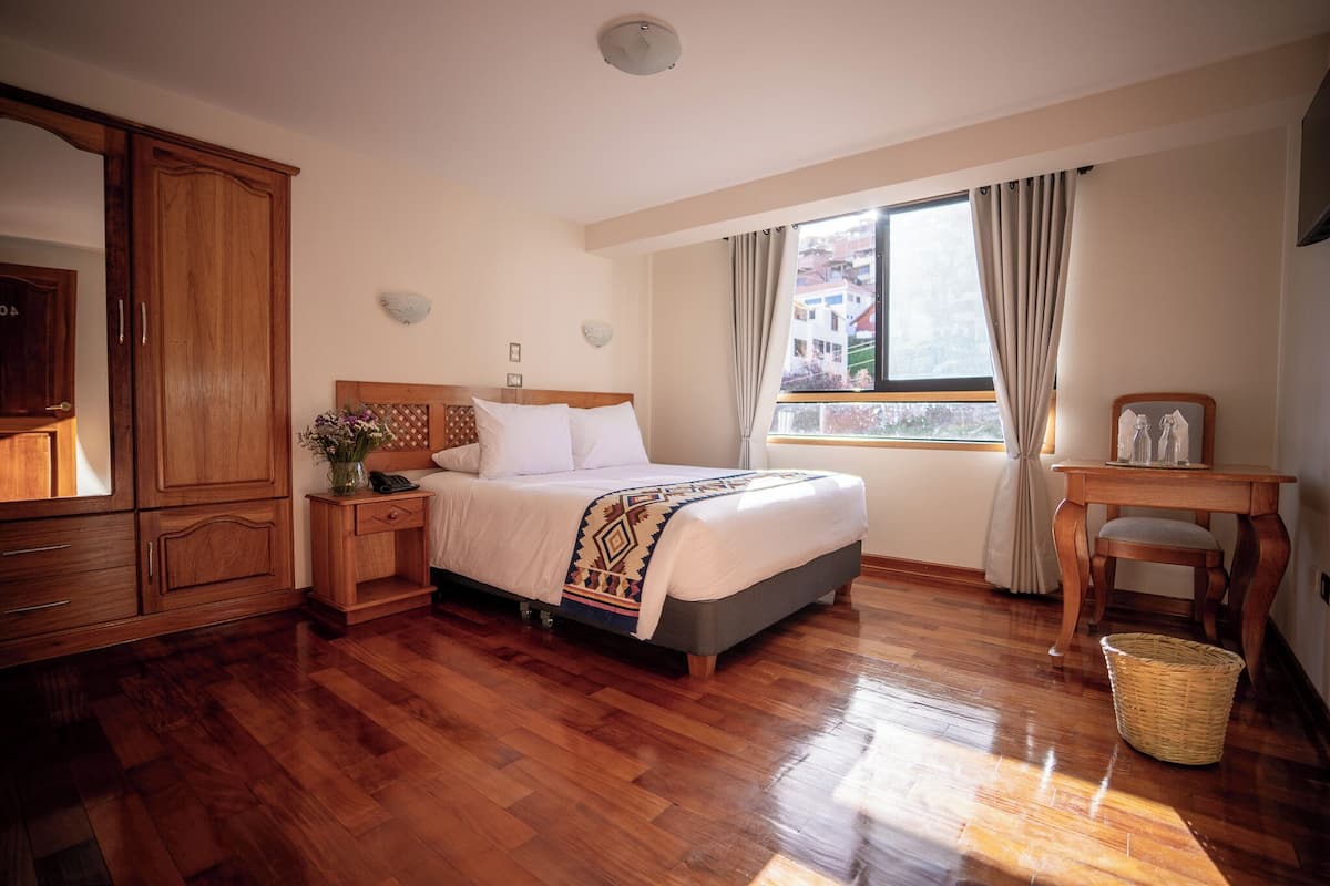 Une chambre d’hôtel avec un lit en bois, une table de chevet en bois surmontée d’un vase, une armoire en bois, une fenêtre avec des rideaux et un panier en osier.